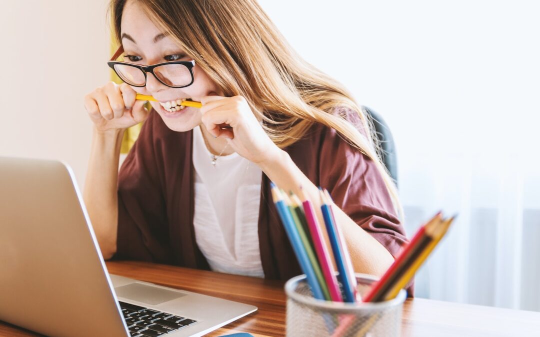 woman biting pencil while sitting on chair in front of computer during daytime